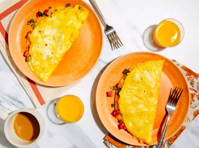 Top-down view of two fluffy omelets served on peach-colored plates, with glasses of orange juice and coffee on the side