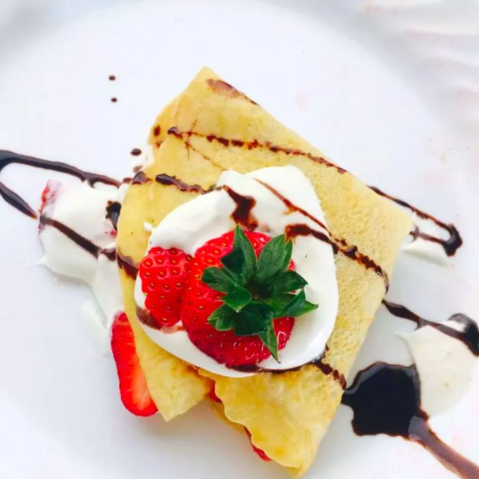 A top-down shot of a crêpe plate, adorned with fresh strawberries, a dollop of whipped cream, and a drizzle of chocolate syrup.