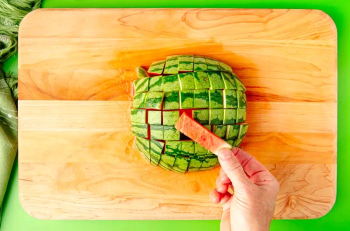 A top-down view of half a watermelon cubed, with a hand pulling out a stick of watermelon with the rind still on top.