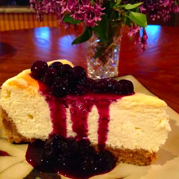 A slice of lemon cheesecake topped with fresh blueberries, with a vase of flowers softly blurred in the background