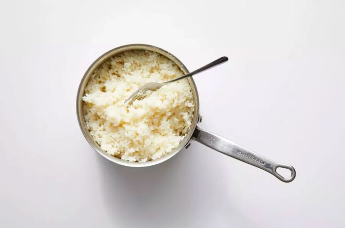 cooked white rice and a fork in a stainless steel KitchenAid saucepan on a white background