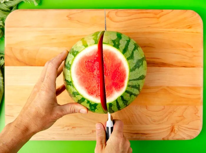 An overhead shot of someone holding a watermelon steady while a knife cuts through its center.