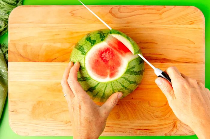 An overhead shot of hands using a knife to peel the rind off a watermelon.