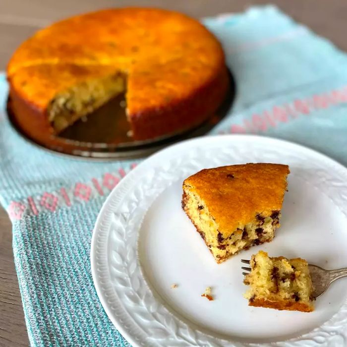 A slice of soft, moist yogurt cake served on a white plate, with the rest of the cake in the background.