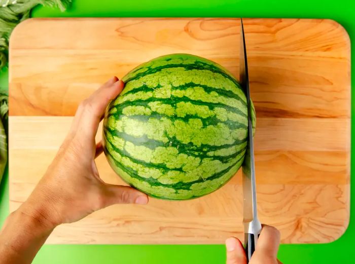 One hand holds the whole watermelon steady on a cutting board, while the other hand cuts off the sides with a sharp chef's knife.