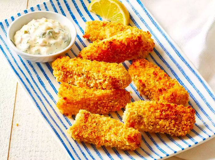 A plate of homemade fish sticks served with tartar sauce on the side, resting on a blue and white striped platter.