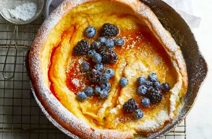 Aerial shot of a Dutch Baby pancake baked in a skillet, adorned with powdered sugar, fresh berries, and drizzled with syrup