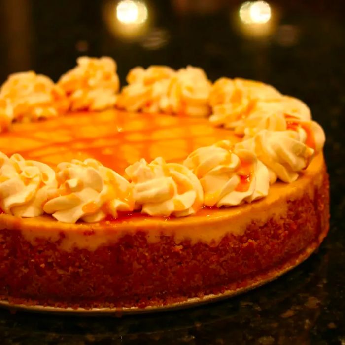 A side view of a smooth, pale brown cheesecake topped with whipped cream rosettes and a sweet caramel drizzle.