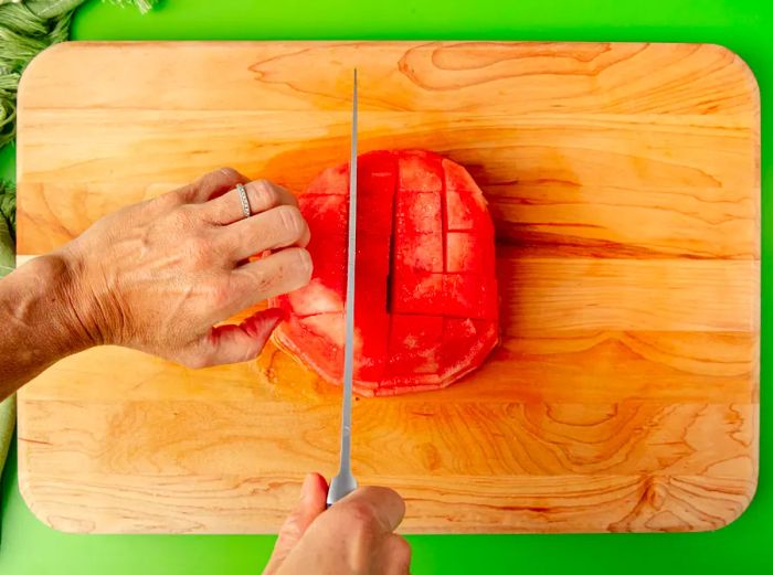 A top-down view of hands cutting the watermelon into cubes by rotating the previously cut sticks.