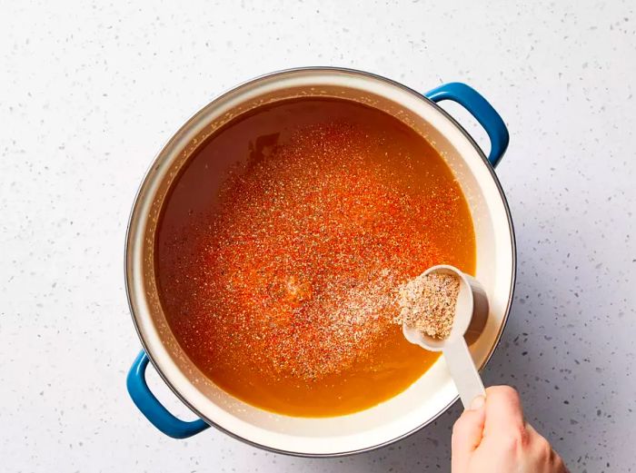 A hand pouring a measured amount of spice blend into a large pot, with the pork shoulder submerged in apple cider vinegar