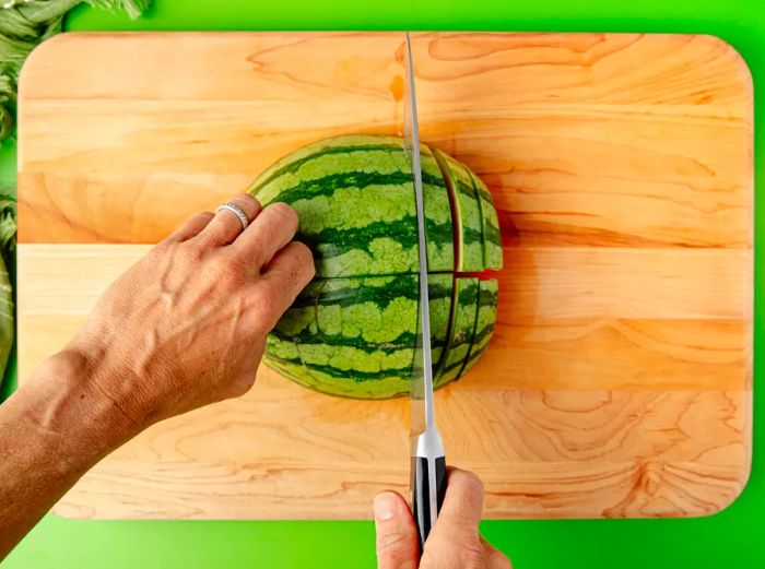 A top-down view of hands cutting two quarters of a watermelon into half-inch thick slices.