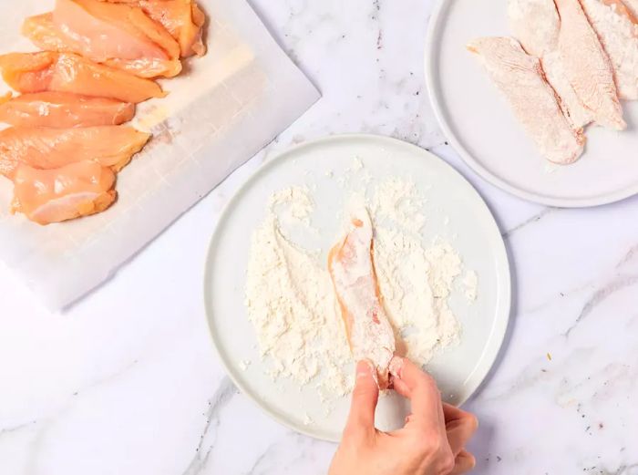 A cutting board displaying thin chicken breast strips, with one being coated in flour.