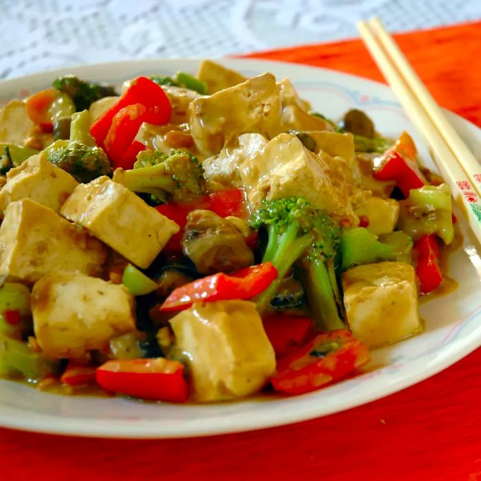 Close-up shot of tofu, broccoli, and peppers drizzled with peanut sauce, served on a plate with chopsticks.