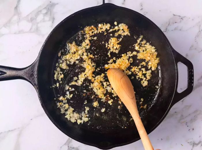 A cast iron skillet with chopped garlic and shallots being stirred with a wooden spoon.