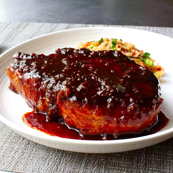 A close-up shot of a Sticky Garlic Pork Chop on a white plate, paired with a rice salad.