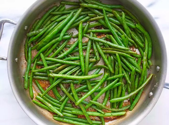 An overhead view of green beans, garlic powder, onion powder, salt, and pepper in a stainless steel pot, cooking over medium-high heat until the beans release moisture.