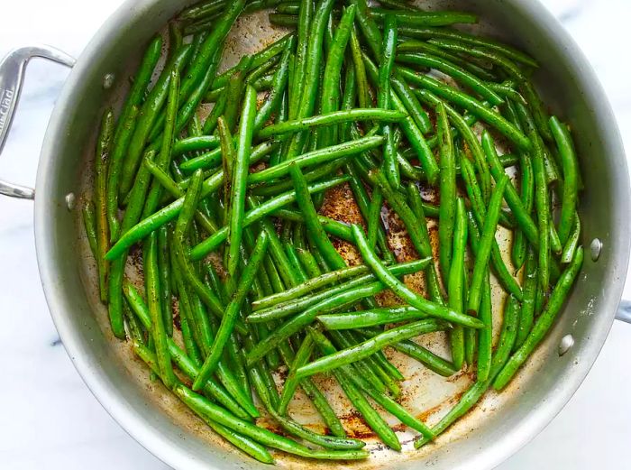 An overhead view of green beans in a large stainless steel pan, slightly charred with olive oil mixed in.