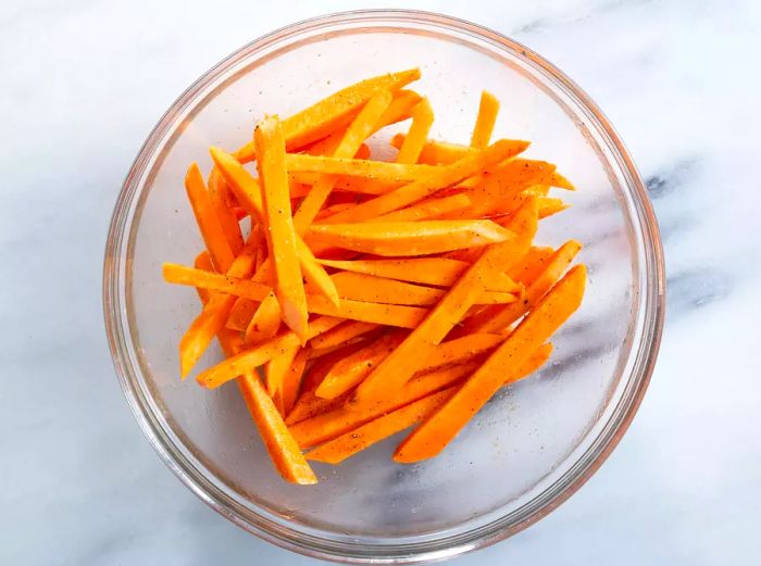 A top-down view of raw sweet potatoes, sliced into fries and seasoned with oil and spices.