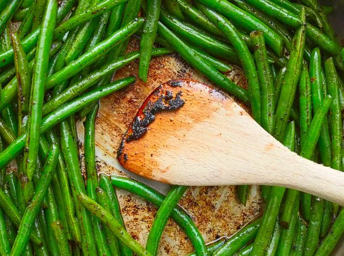 An overhead view of the bottom of a stainless steel pan with green beans being scraped to incorporate the charred spices.