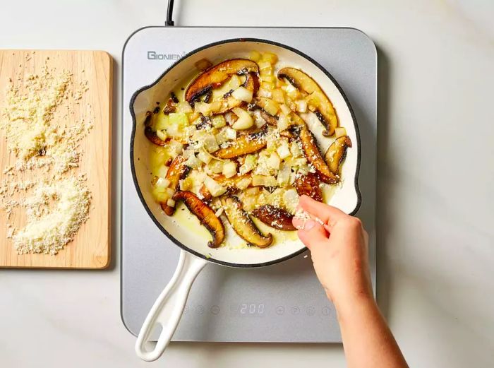 A hand sprinkling grated cheese over sautéed mushrooms and onions in a pan.