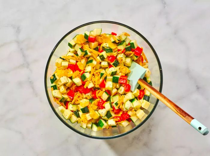 A large glass mixing bowl filled with zucchini, bell peppers, cornflakes, cheese, oil, basil, and eggs