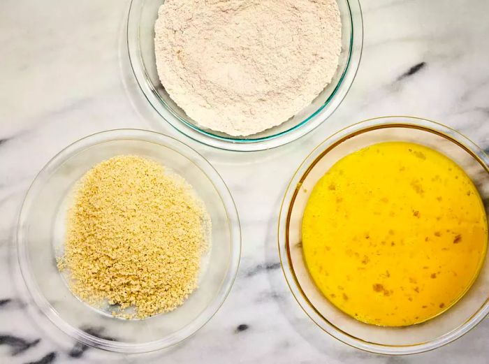 A breading station set up with three bowls: one containing seasoned flour, another with beaten eggs, and the third with panko breadcrumbs.