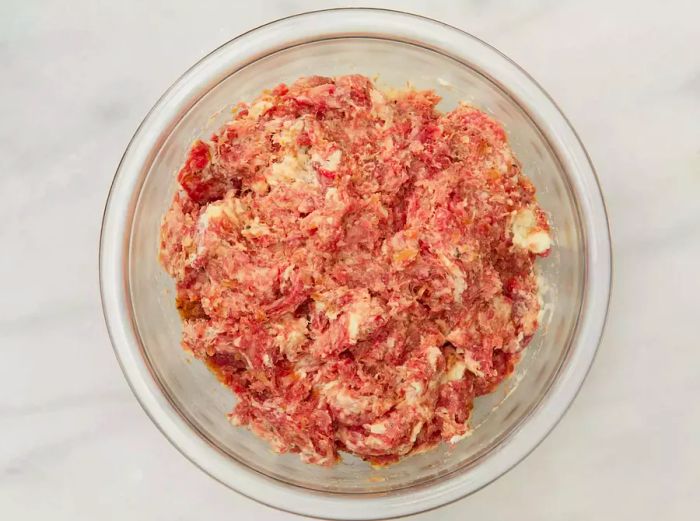 Aerial view of a bowl with ground beef, pork, egg, brown sugar, salt, black pepper, nutmeg, allspice, ginger, and onions for the Swedish Meatballs (Svenska Köttbullar) recipe.