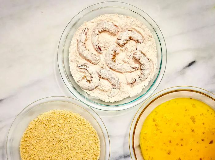 A bowl of seasoned flour with shrimp being coated, alongside bowls of panko breadcrumbs and whisked eggs.