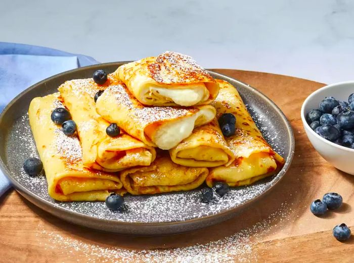 A plate of golden-brown cheese blintzes, sprinkled with powdered sugar and topped with fresh blueberries.