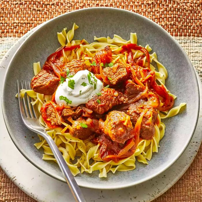 A close-up, overhead shot of a single serving of Hungarian goulash, served atop egg noodles and topped with a dollop of sour cream.