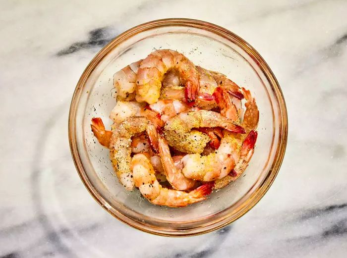 A clear bowl containing raw shrimp seasoned with salt, pepper, and garlic powder.