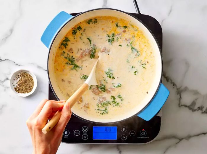 A pot of sausage, potato, and kale soup being stirred with a wooden spoon