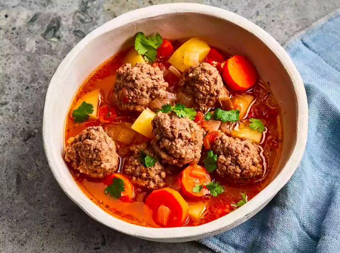 Aerial view of a large bowl filled with albondigas, featuring meatballs, carrots, potatoes, fresh herbs, and broth.