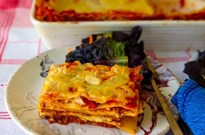 A close-up shot of a perfectly plated slice of lasagna, resting in front of the baking pan filled with more lasagna.