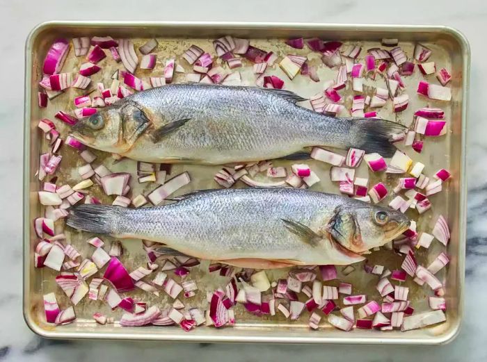 A bird's-eye view of two whole branzino fish placed on a sheet pan with red onions.