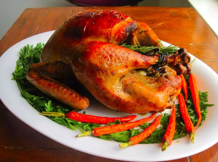 A beautifully roasted turkey on a white platter, garnished with fresh herbs and whole roasted carrots.