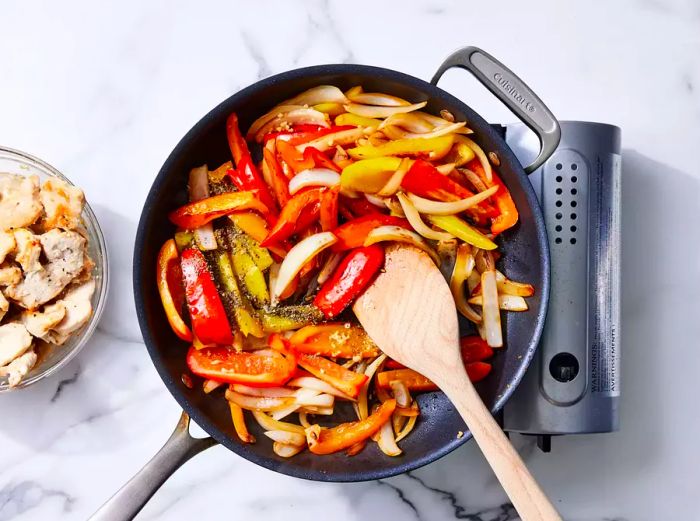 A pan with sliced peppers, onions, garlic, basil, and balsamic vinegar being stirred with a wooden spoon