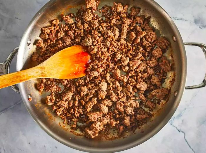 Ground beef browning in a skillet with a wooden spoon.