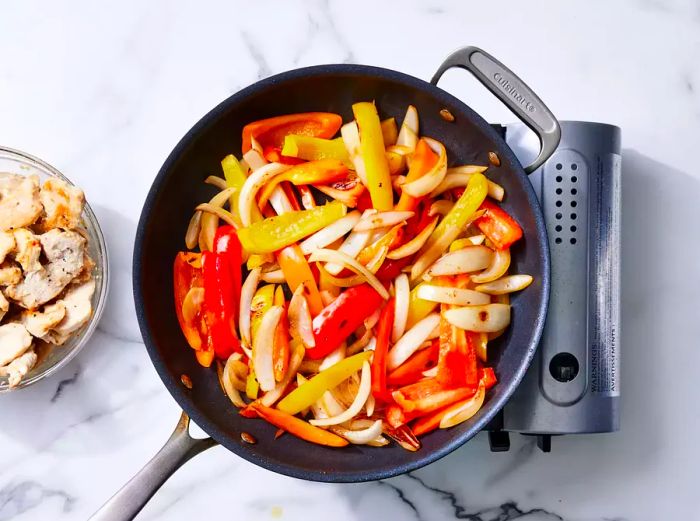 A large skillet filled with sliced peppers and onions, accompanied by a small bowl of cooked chicken on the side