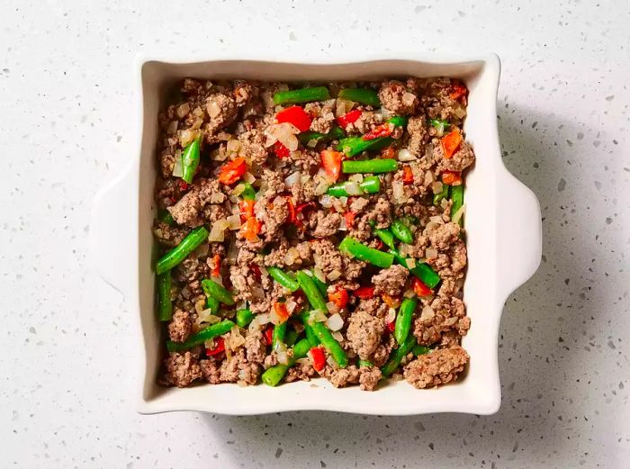 A greased baking dish filled with cooked ground beef, green beans, onions, and tomatoes.