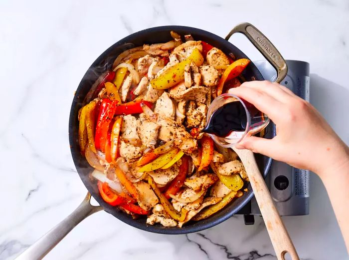 A hand pouring balsamic vinegar from a small measuring cup into a pan with chicken, peppers, and onions.