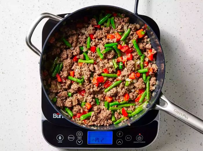 A skillet with browned ground beef, green beans, tomatoes, onions, and garlic all together.