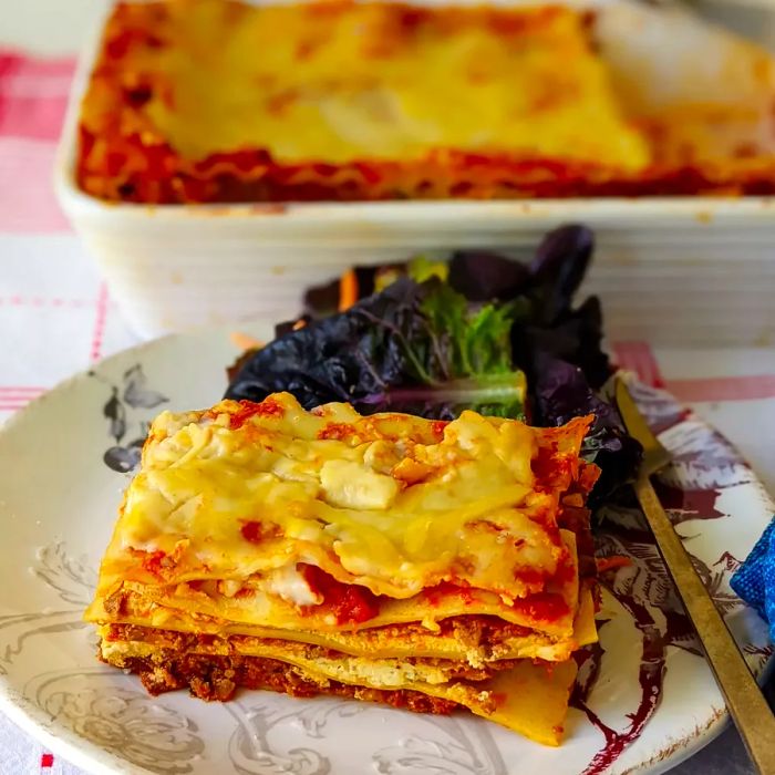 A close-up shot from a low angle of a beautifully plated slice of lasagna in front of the full pan of lasagna.