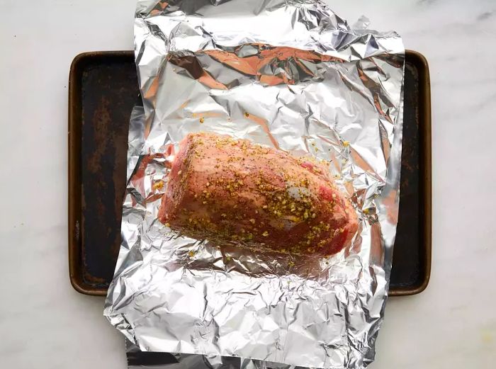 A raw beef roast coated with seasoned paste, resting on tinfoil on a baking sheet.