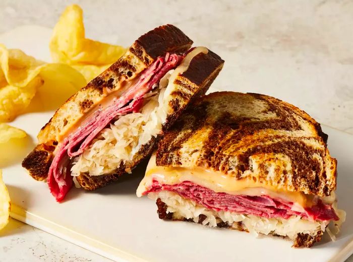 A Reuben sandwich cut in half, served alongside crispy potato chips