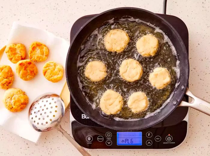 A plate lined with paper towels holding Jamaican fried dumplings, next to a pan with more dumplings frying in hot oil.