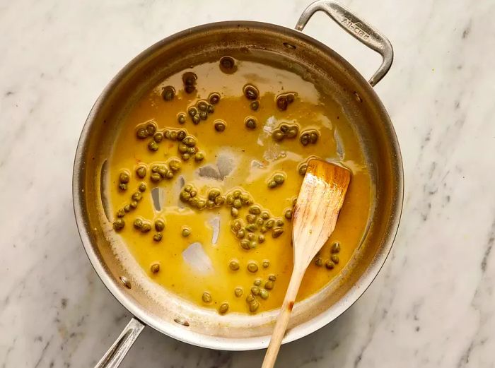 Lemon-caper sauce simmering in a pan, being stirred with a wooden spoon, viewed from above