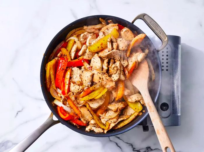 A pan with seasoned chicken, peppers, and onions being stirred with a wooden spoon