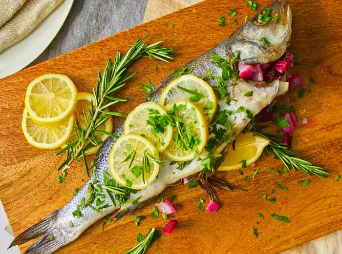 A top-down view of Branzino Mediterranean garnished with lemon slices and sprigs of rosemary on a cutting board.