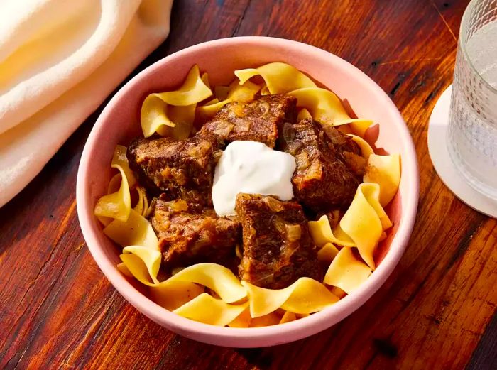 Close-up of a bowl of egg noodles topped with hearty beef goulash and a dollop of sour cream.